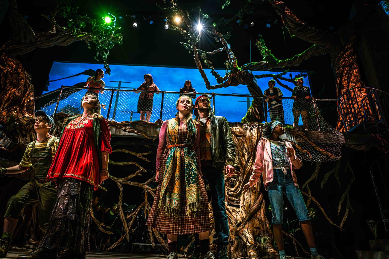 A dramatic stage production featuring a group of diverse actors looking upward with expressions of awe and anticipation. The set design is highly detailed, featuring massive, twisted artificial trees with textured bark, a chain-link fence on a raised platform, and a blue-sky backdrop. Bright green and white spotlights illuminate the performers from above.