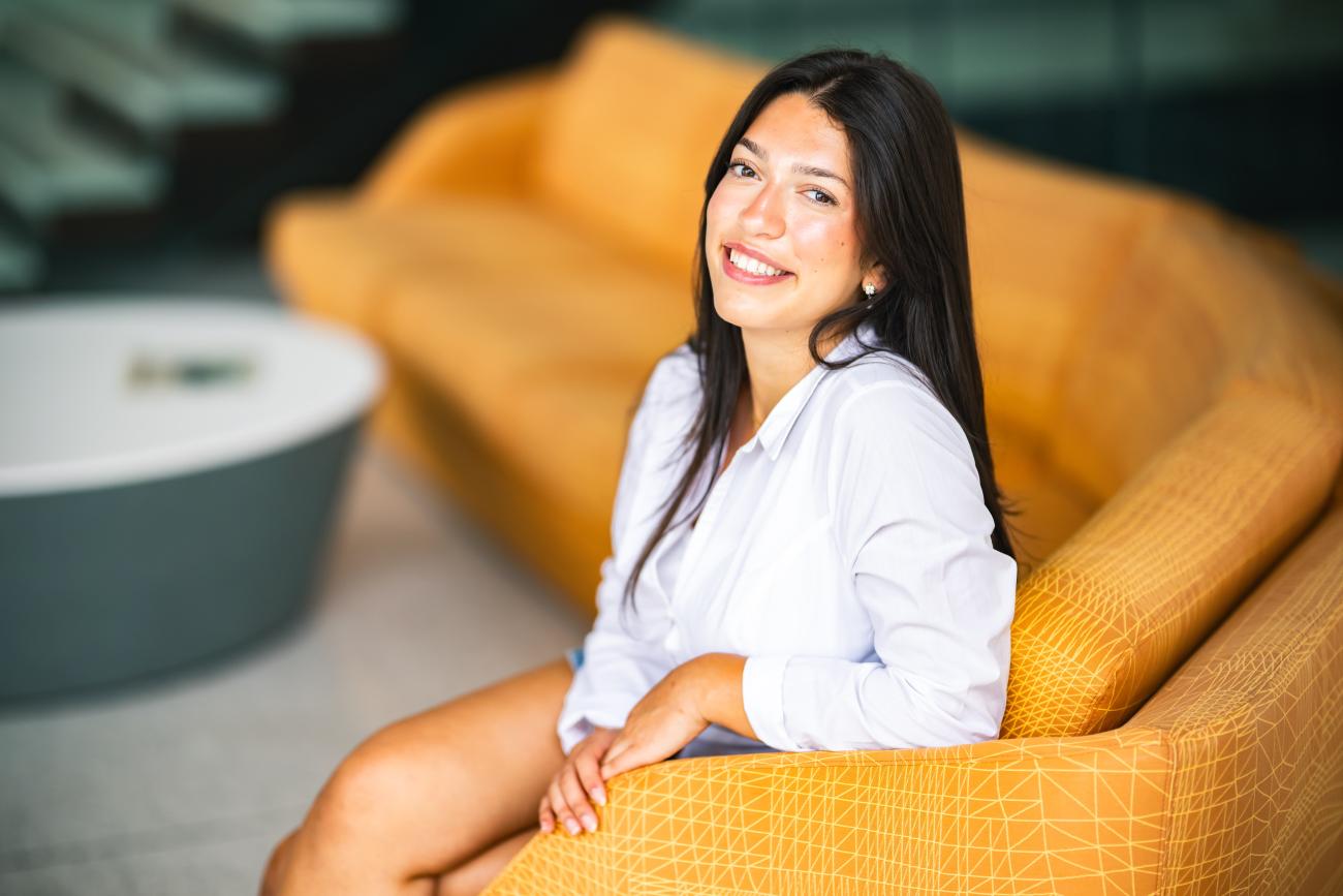 Jordan Kotch sits on an orange sofa and smiles at the camera wearing a white button up shirt.